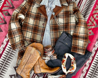 May include: A brown and white plaid jacket with a fur collar, a white button-down shirt with red stripes, a pair of dark gray pants, a pair of brown cowboy boots, and a white and orange bandana. The jacket has the word "PLAINS" on the pocket.