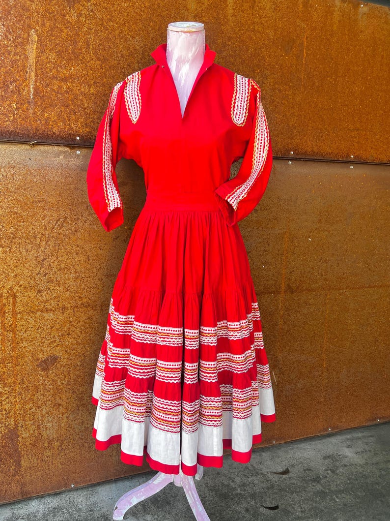 May include: A red vintage dress with white and gold embroidery. The dress has a full skirt with multiple layers of white and red trim. The dress is on a white mannequin.
