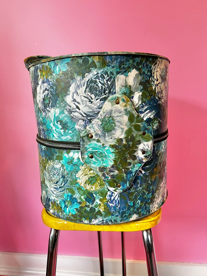 May include: A vintage floral patterned hat box with a zipper closure and a metal accent on the side. The hat box is sitting on a yellow stool.