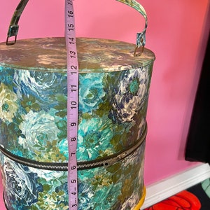 May include: A vintage hat box with a floral print in shades of blue, green, and white. The box has a zipper closure and a handle. The box is 17 inches tall.