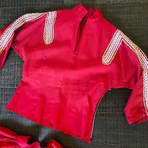 May include: A red blouse with white lace trim on the sleeves and neckline. The blouse has a V-neck and a zipper closure in the back.