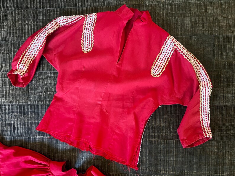 May include: A red blouse with white lace trim on the sleeves and neckline. The blouse has a V-neck and a zipper closure in the back.