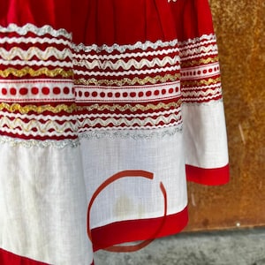 May include: A red skirt with white and gold trim. The skirt has a white cotton lining and a red waistband. The skirt is made of a lightweight fabric and has a gathered design.