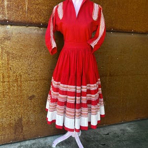 May include: A red vintage dress with white and gold embroidery. The dress has a full skirt with multiple layers and a fitted bodice. The dress is on a white mannequin.