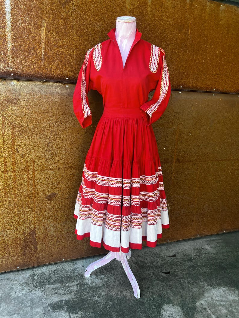 May include: A red vintage dress with white and gold embroidery. The dress has a full skirt with multiple layers and a fitted bodice. The dress is on a white mannequin.