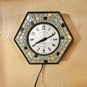 Puede incluir: Un reloj de pared General Electric vintage con un marco hexagonal de mosaico. La esfera del reloj es blanca con números y manecillas negras. Los azulejos de mosaico son en tonos de gris, negro, amarillo y colores pastel. Un cable de alimentación negro cuelga debajo.