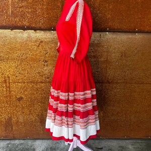 May include: A red dress with white and orange embroidery. The dress has a high neck and a full skirt. The skirt is tiered and has a white lace trim.
