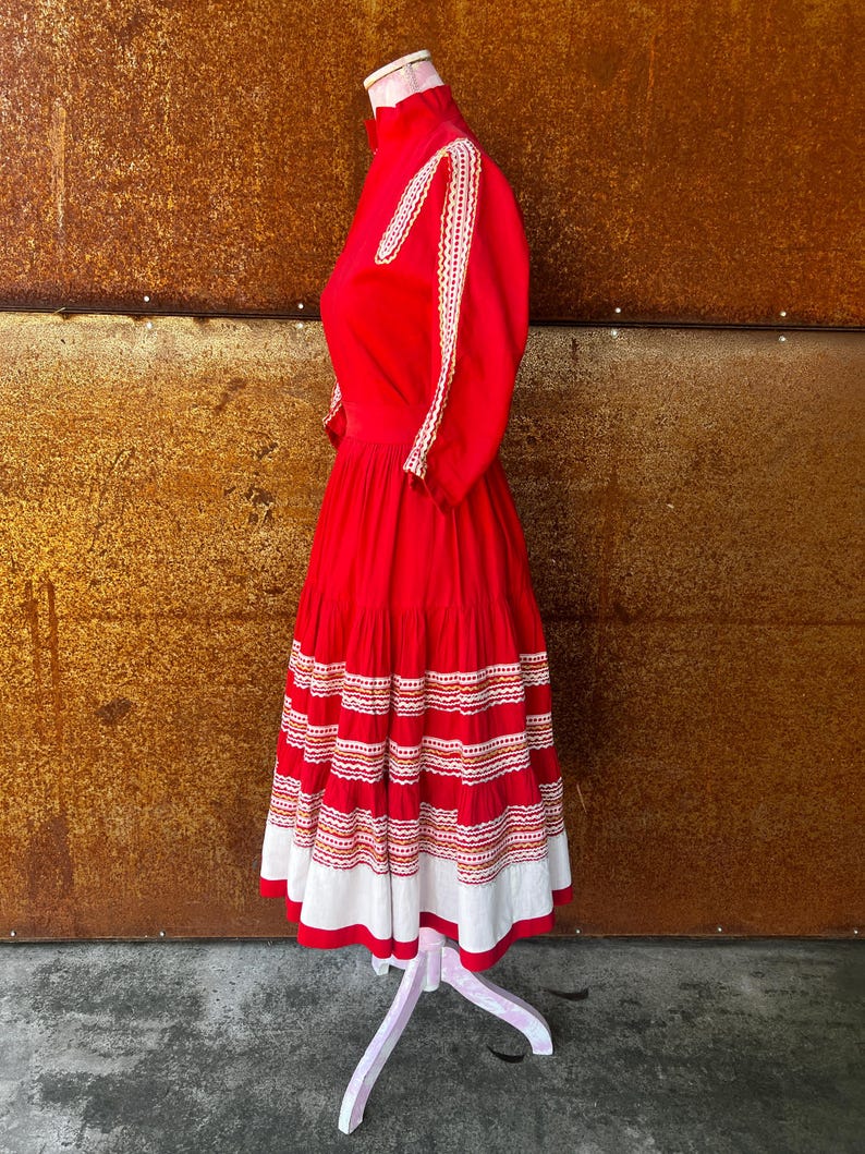 May include: A red dress with white and orange embroidery. The dress has a high neck and a full skirt. The skirt is tiered and has a white lace trim.