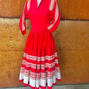 May include: A red dress with white and orange embroidery. The dress has a fitted bodice and a full skirt with multiple tiers. The sleeves are three-quarter length and have embroidery on the shoulders.