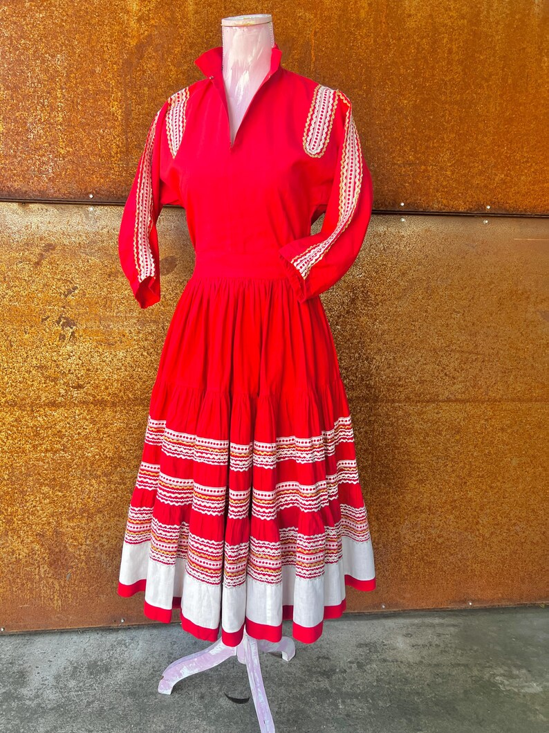 May include: A red dress with white and orange embroidery. The dress has a fitted bodice and a full skirt with multiple tiers. The sleeves are three-quarter length and have embroidery on the shoulders.
