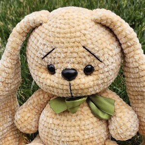 May include: A plush, beige bunny rabbit with long floppy ears and a green ribbon bow. The handmade stuffed animal has black eyes and a black nose. The bunny is sitting on green grass.
