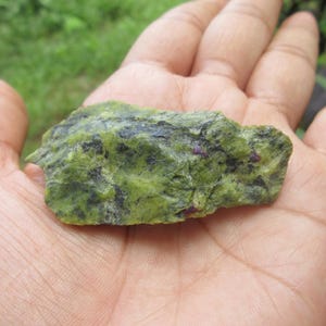 May include: A rough green stone with dark patches and small red inclusions. The stone is held in an open hand, with a blurred green background. The stone's texture is uneven, with a natural, raw appearance.