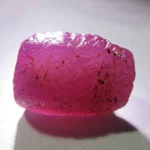 May include: A raw, uncut ruby gemstone with a vibrant pink hue. The surface of the stone has a rough, textured appearance with small dark inclusions. The gemstone is displayed against a white background, showcasing its natural form and color.