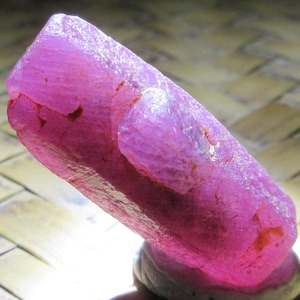 May include: A raw, elongated crystal specimen with a vibrant, translucent magenta-pink hue. The stone features a faceted, irregular surface with visible internal fractures, natural crystalline textures, and small patches of earthy brown inclusions.