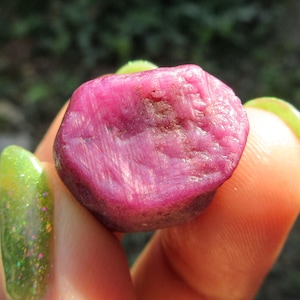 May include: A large, faceted, pink ruby gemstone with a rough, natural finish. The ruby is held in a hand, showing its size and shape.