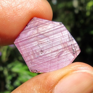 May include: A faceted, hexagonal-shaped, pink gemstone with parallel striations. The gemstone is held between two fingers, with a blurred green background.