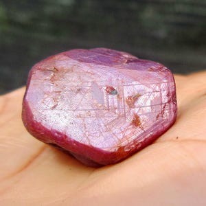 Könnte beinhalten: Ein roher, ungeschliffener Rubin-Edelstein mit tiefroter Farbe und leicht durchscheinendem Aussehen. Die Oberfläche des Steins weist sichtbare Linien und Unvollkommenheiten auf, was ihm ein natürliches, unpoliertes Aussehen verleiht. Der Rubin wird in einer Hand gehalten.