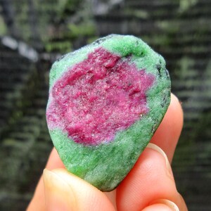 May include: A natural ruby in zoisite gemstone. The stone features a vibrant pinkish-red center surrounded by a green outer layer. The rough, uncut gemstone is held in a hand, showcasing its natural texture and color variations.