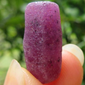 Puede incluir: Una piedra preciosa de rubí en bruto de forma rectangular, con un tono magenta rosado intenso y una superficie texturizada mate. La piedra presenta inclusiones oscuras naturales dispersas, mostrando una forma cristalina alargada.