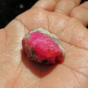 Könnte beinhalten: Ein roher, rosafarbener Rubin-Edelstein mit einer weißen, trüben Außenseite. Der Rubin liegt in der Handfläche.