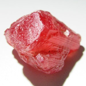 Puede incluir: Primer plano de una gema roja en bruto sin cortar. La gema tiene una estructura cristalina con líneas y facetas visibles. La piedra es de un tono rojo vibrante, con una apariencia ligeramente translúcida. La gema está sobre un fondo blanco.