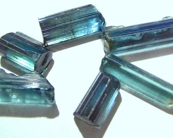 Puede incluir: Un conjunto de cristales de turmalina en bruto, de color azul verdoso. Los cristales son alargados, con una textura estriada y diferentes tonos de azul y verde. Están dispersos sobre una superficie blanca, mostrando su belleza natural.