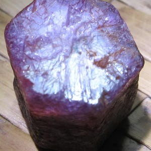 Puede incluir: Un cristal de rubí en bruto de forma hexagonal con un tono magenta y púrpura intenso. La superficie superior muestra una textura cristalina compleja con fracturas internas visibles y facetas que reflejan la luz, mientras que los lados son opacos.