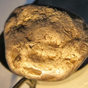 May include: A close-up of a gold nugget with a rough, textured surface. The nugget has a rounded shape and varying shades of gold, with darker areas suggesting shadows or imperfections. The lighting highlights the metallic luster of the gold.