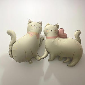 May include: Two beige cat-shaped balloons. One cat has a pink bow tie, and the other has a pink heart. Both have closed eyes and a pink collar. The balloons have small gold star accents.