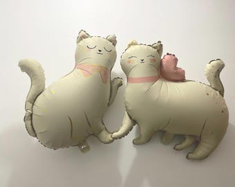 May include: Two beige cat-shaped balloons. One cat has a pink bow tie, and the other has a pink heart. Both have closed eyes and a pink collar. The balloons have small gold star accents.