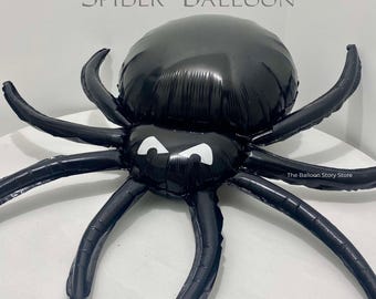 May include: A black inflatable spider balloon with white eyes. The balloon is shaped like a spider with eight legs and a large body. The balloon has the text "The Balloon Story Store" printed on it.