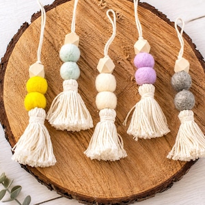 May include: Five hanging ornaments featuring cream-colored cotton tassels, natural wooden geometric beads, and two felt balls in yellow, light blue, cream, lavender, and gray. Each ornament includes a white cord loop for hanging purposes.
