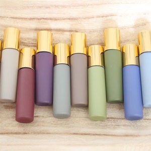 Puede incluir: Un surtido de diez frascos roll-on en varios colores pastel, incluyendo lavanda, gris, burdeos y verde. Cada frasco tiene una tapa dorada. Los frascos están dispuestos sobre una superficie de madera clara.