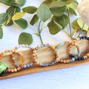 May include: A wooden tray holds several beaded bracelets with various stone and wood beads. An essential oil bottle with a green label and the words "Stress Away" is next to the bracelets. Green foliage and a white crystal are in the background.