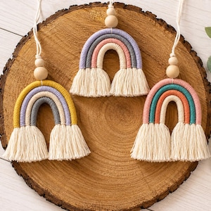 Puede incluir: Tres adornos colgantes de arcoíris en macramé con cordón de algodón envuelto en colores amarillo, gris, lavanda, rosa, verde azulado y naranja. Cada arcoíris tiene una cuenta de madera natural en la parte superior y flecos color crema.