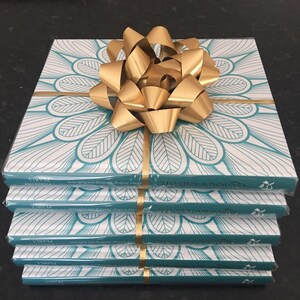 May include: A stack of four books wrapped in clear plastic, with a teal and white floral design on the covers. A large gold bow sits on top. Gold ribbon wraps around the books. The book title is visible.