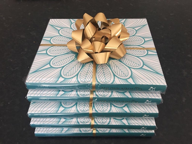 May include: A stack of four books wrapped in clear plastic, with a teal and white floral design on the covers. A large gold bow sits on top. Gold ribbon wraps around the books. The book title is visible.