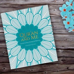 May include: A book titled "Qur'an and Me" with a teal and white floral design on the cover. The book's title is in gold lettering, and the cover features a large teal flower shape. The book is on a wooden surface with blue paper flowers.