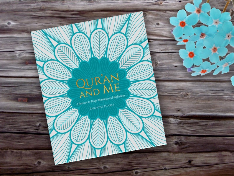 May include: A book titled "Qur'an and Me" with a teal and white floral design on the cover. The book's title is in gold lettering, and the cover features a large teal flower shape. The book is on a wooden surface with blue paper flowers.