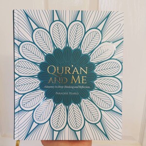 May include: A book titled "Qur'an and Me" with a teal and white floral design. The title is in gold lettering, with the subtitle "A Journey to Deep Thinking and Reflection." The book is held by a person.