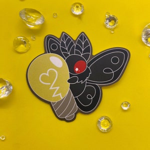 May include: A black and red moth with a light bulb in its mouth. The moth is on a yellow background with small, clear rhinestones.