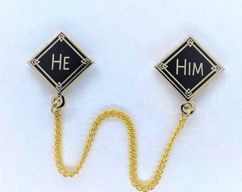 May include: Two black diamond-shaped pins with gold trim and a gold chain connecting them. The pins say "HE" and "HIM" in gold lettering.