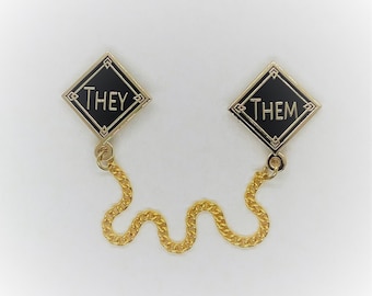 May include: Two black enamel pins with gold trim, one says "THEY" and the other says "THEM". The pins are connected by a gold chain.