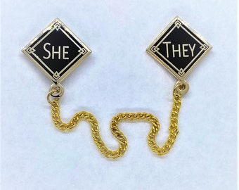 May include: Two black diamond-shaped pins with gold trim and a gold chain connecting them. The pins say "SHE" and "THEY" in white lettering.