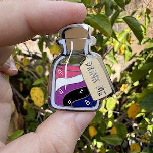 Könnte beinhalten: Ein silberfarbener Emaille-Pin in Form einer Zaubertrankflasche. Die Flasche enthält Schichten aus Rosa, Weiß, Schwarz, Lila und Blau. Ein Etikett auf der Flasche trägt die Aufschrift "DRINK ME". Der Pin wird von einer Hand gehalten.