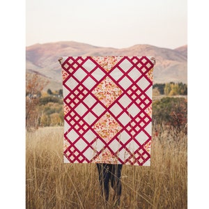 Pode incluir: Uma pessoa segura uma colcha vermelha e branca com um padrão geométrico. A colcha está em um campo de grama alta. O texto "Modern Chalet Quilt | Throw | Baby |" está no topo da imagem. O texto "Taren Studios" está na parte inferior da imagem.