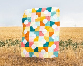 Puede incluir: Una colcha colorida con un patrón geométrico de cuadrados y círculos en tonos de rosa, amarillo, turquesa y blanco. La colcha se muestra en un campo de hierba alta.