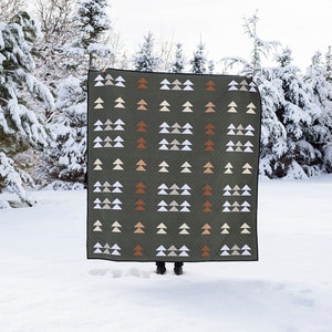 May include: A green quilt with a geometric pattern of white and orange triangles, resembling stylized trees. The quilt is displayed outdoors in a snowy setting.