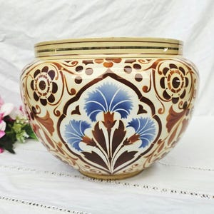 May include: A ceramic planter with a cream base and detailed floral designs in brown, blue, and cream. The planter has a wide, rounded form and a slightly flared rim, suitable for displaying plants. A decorative piece.