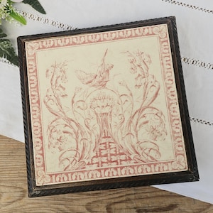 Puede incluir: Obra de arte cuadrada enmarcada con un fondo crema y un diseño floral y de pájaros rojo. El marco marrón oscuro tiene una textura de cuerda. La obra de arte se exhibe sobre una superficie de madera.